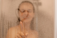 Photo Of Smiling Attractive Young Woman Draws Heart On Weeping Glass Shower Door, Enjoys Rest In Douche, Washing Her Body, Standing Behind Steam Blurred Glass With Water Drops.