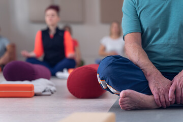 Detail of hands and feet with wrinkles, of an elderly man, sitting in lotus position, doing yoga with other people.