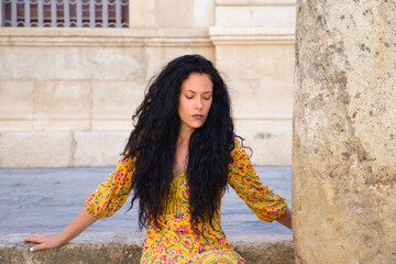 Spanish woman with curly brown hair and beautiful, is sitting posing for the camera on the steps of a staircase and next to a column. tourism and travel concept.