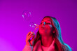 © master1305 - Young beautiful girl with cute freckles in white sweater blowing soap bubbles isolated on purple color studio background in neon light.