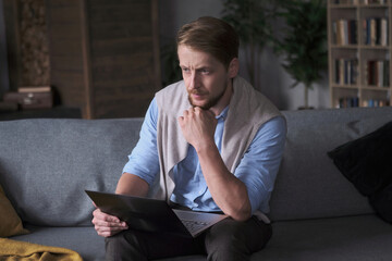 Confident European business male working remotely use laptop on couch at cozy living room. Modern businessman surfing internet distance chatting on computer digital technology at home apartment