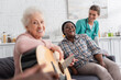 © LIGHTFIELD STUDIOS - Positive interracial women with acoustic guitar looking at camera near nurse in nursing home