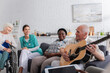 © LIGHTFIELD STUDIOS - Senior man holding acoustic guitar near multiethnic friends and nurse in nursing home