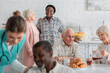 Senior woman holding teapot near friend and blurred interracial people in nursing home