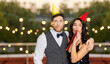 © Syda Productions - birthday, celebration and holidays concept - happy couple with party blowers and caps having fun at rooftop party over lights on background