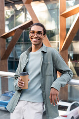 Wall Mural - pleased african american man in eyeglasses holding coffee to go while smiling at camera outdoors