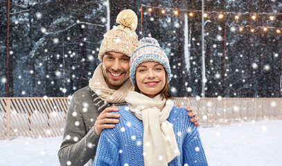 Canvas Print - people, christmas and winter holidays concept - happy couple in knitted hats and scarves over ice rink background