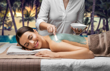 Poster - wellness, beauty and cosmetology concept - beautiful young woman lying with closed eyes and cosmetologist applying clay mask by spatula at spa over tropical beach background in french polynesia