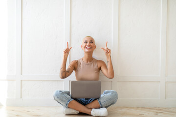 Wall Mural - Check this. Excited young lady sitting with laptop and pointing up at free space above her head over white wall