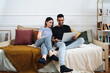 © diignat - Happy young mixed race couple enjoying time at home using computer sitting on sofa, black man and white woman having fun online shopping or surfing web relaxing on couch