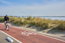 Bicycle Path In The Netherlands. Dune With Grass In Front Of A Blue Sea.1 Male Bicyclist Motion Blurred. Blue Sky For Text Free Space. In South Holland On The Brouwersdam At The Grevelinger Sea.