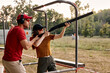 © Roman - young man and woman in goggles and headset preparing to shoot, ready to shoot. Skilled experienced man is teaching female in outdoor range. Firearms for sports shooting, hobby. Side view.
