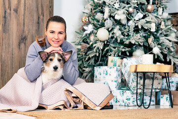 Happy woman with her dog greets, merry Christmas and New Year. The female sits with her pet by the Christmas tree