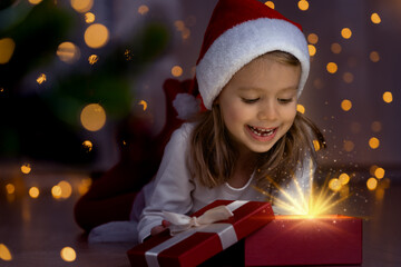 Wall Mural - Close up of little girl at dark in red santa hat opens box with magic light and laughs next to christmas tree. Happy child with New Year gift