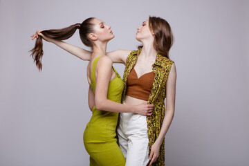 Two high fashion models in robe-dress, white pants, brown top, shoes, yellow sage gold dress. Beautiful young women. Studio shot, portrait. White, soft gray background.