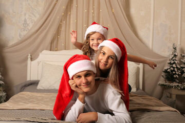 Wall Mural - a young family with a little daughter in Santa Claus hats are lying on the bed at home Christmas