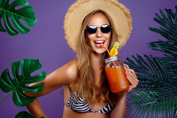 Wall Mural - Cheerful girl drinks fruit cocktail in sunglasses and straw hat while stands near tropical plants 