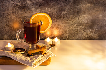 Wall Mural - mulled wine with orange slice on wooden plate with towel on white ang dark background. led light candle 