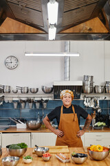 Wall Mural - Vertical portrait of smiling female cook looking at camera while posing in industrial kitchen interior, copy space