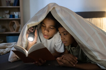 Wall Mural - African American girl reading book to younger brother under light of flashlight while they lying under duvet in dark room