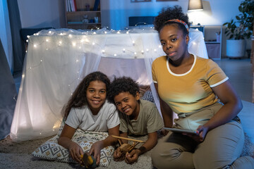 Canvas Print - Portrait of careful African American mother with tablet sitting at handmade tent and reading fairy tale to kids