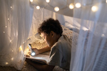 Curious Black Boy With Curly Hair Lying Inside Of Play Tent Decorated With Christmas Lights And Reading Book With Flashlight
