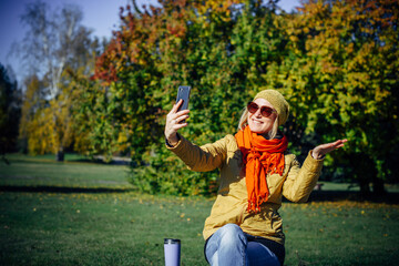 Wall Mural - Cheerful girl in bright clothes and sunglasses sitting on park bench and taking selfie on smartphone against autumn trees. Attractive woman makes hand gestures on the background of nature. Copy space.