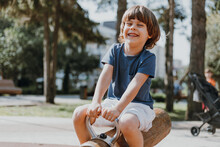 Happy Laughing Little Boy In A Blue T-shirt And White Shorts Rides On A Wooden Rocking Chair Outdoor. Child Is Playing On A Public Playground On A Sunny Summer Day. Lifestyle. Space For Text