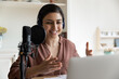 Smiling young indian female radio program host or blogger in headphones recording audio or voice acting using professional stand microphone, communicating in social network or streaming online.