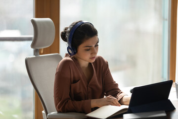 Canvas Print - Concentrated young indian ethnicity business woman in wireless headphones holding distant video call meeting using digital computer tablet, writing notes in paper planner or studying on online courses