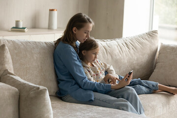 Wall Mural - Happy beautiful millennial mother embracing sincere cute kid daughter, learning using software cellphone applications, web surfing information, shopping in internet store, resting on cozy sofa.