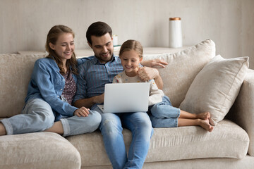 Wall Mural - Happy young Caucasian couple parents using computer applications with laughing small cute kid daughter, enjoying playing online games, choosing goods shopping in internet store, watching cartoons.