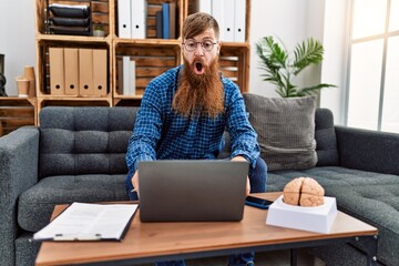 Canvas Print - Redhead man with long beard using laptop working at psychology clinic scared and amazed with open mouth for surprise, disbelief face