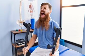 Sticker - Redhead man with long beard holding crutches at rehabilitation clinic angry and mad screaming frustrated and furious, shouting with anger. rage and aggressive concept.