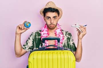 Wall Mural - Young hispanic man wearing summer style and hawaiian lei holding world ball and plane toy depressed and worry for distress, crying angry and afraid. sad expression.