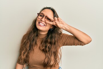 Wall Mural - Young hispanic girl wearing casual clothes and glasses doing peace symbol with fingers over face, smiling cheerful showing victory