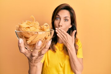 Sticker - Young latin woman holding bowl with uncooked pasta covering mouth with hand, shocked and afraid for mistake. surprised expression