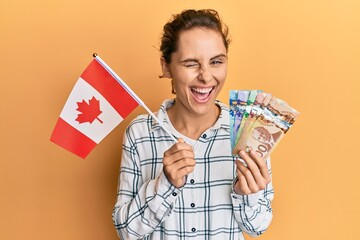 Sticker - Young brunette woman holding canada flag and dollars winking looking at the camera with sexy expression, cheerful and happy face.