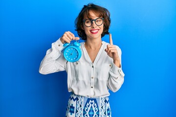 Sticker - Young brunette woman holding alarm clock smiling with an idea or question pointing finger with happy face, number one