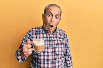 Poster - Handsome senior man with grey hair drinking a cup coffee in shock face, looking skeptical and sarcastic, surprised with open mouth