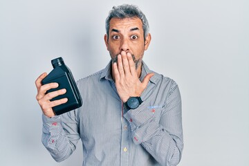 Poster - Handsome middle age man with grey hair holding motor oil bottle covering mouth with hand, shocked and afraid for mistake. surprised expression