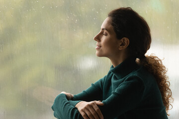 music of rain. side shot of serene latin woman relax by window with closed eyes listen sound of rain