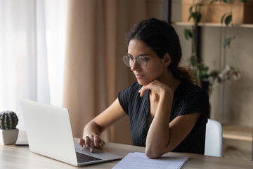 Interesting task. Focused young hispanic woman in eyewear sit by desk at home work online by laptop pc. Millennial female office worker employee search information at corporate network read e document