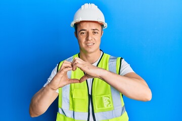 Handsome young man wearing safety helmet and reflective jacket smiling in love showing heart symbol and shape with hands. romantic concept.