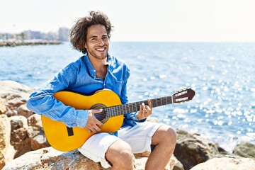 Canvas Print - Young hispanic man playing classical guitar sitting on rock at the beach.