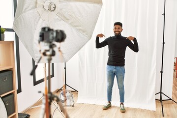 Wall Mural - Young hispanic man with beard posing as model at photography studio looking confident with smile on face, pointing oneself with fingers proud and happy.