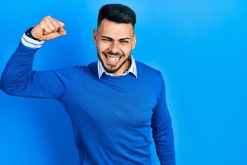 Wall Mural - Young hispanic man with beard wearing casual blue sweater angry and mad raising fist frustrated and furious while shouting with anger. rage and aggressive concept.