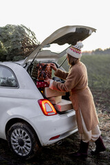 Wall Mural - Woman packing gifts into the car with Christmas tree on a rooftop on nature at dusk. Getting ready for a New Year holidays. Idea of a Christmas mood. Woman wearing fur coat and hat