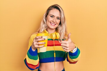 Sticker - Beautiful young blonde woman drinking cup of coffee wearing headphones approving doing positive gesture with hand, thumbs up smiling and happy for success. winner gesture.