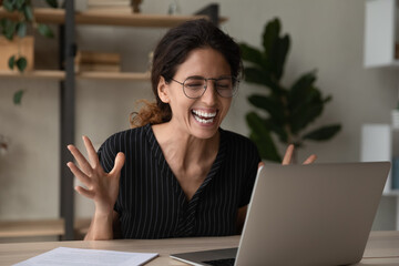 Wall Mural - Overjoyed young casual hispanic woman employee in glasses having fun chat with colleague on laptop in corporate web conference app. Laughing millennial female worker watch funny video at social media